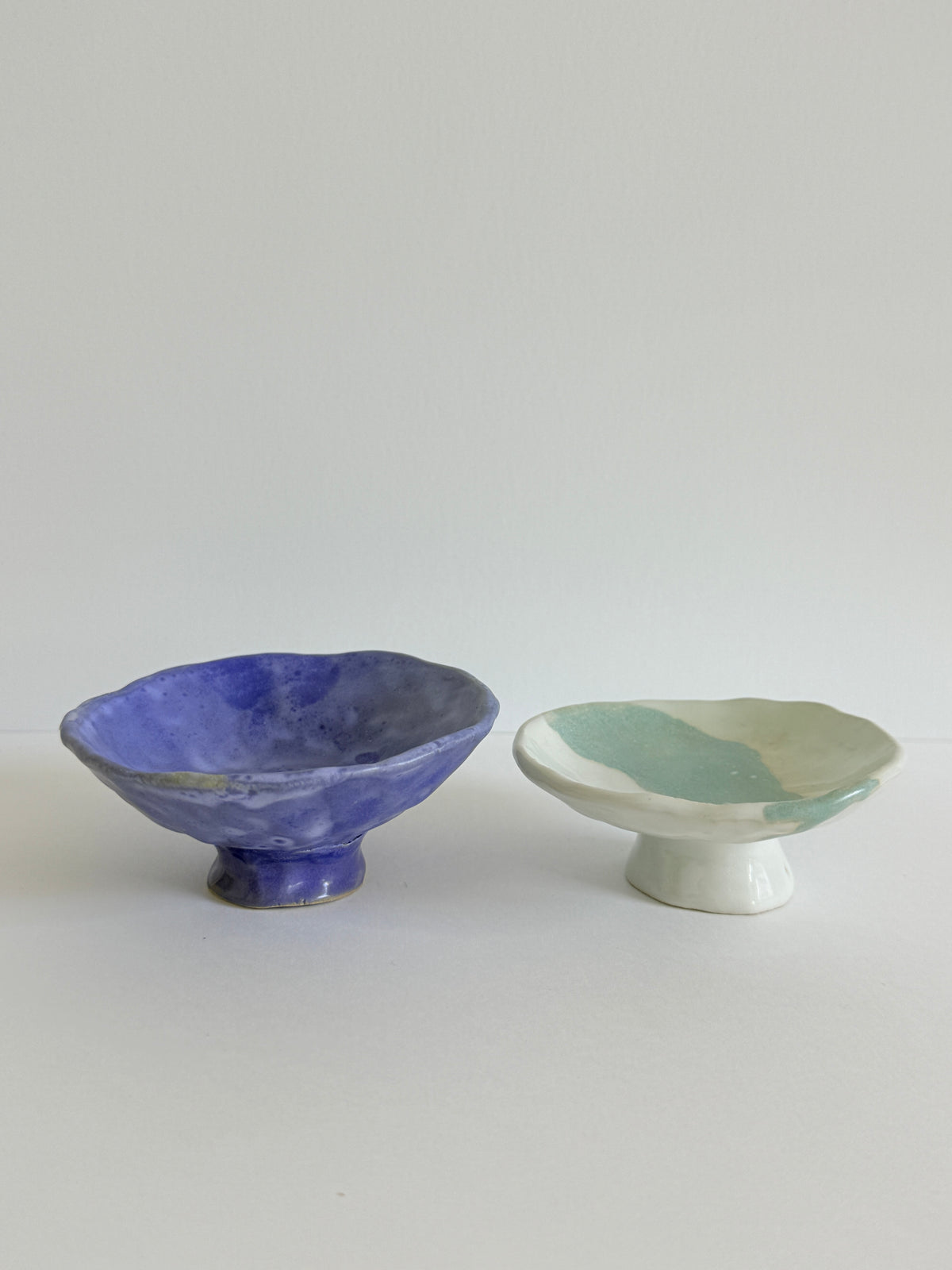 Two handmade ceramic pedestal bowls on a light gray background. The bowl on the left is glazed in a textured cobalt blue, while the bowl on the right is glazed with a marbled pattern of mint green and off-white.
