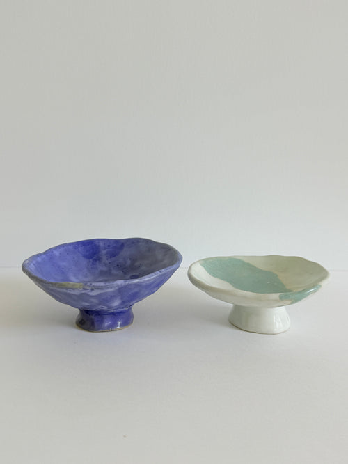 Two handmade ceramic pedestal bowls on a light gray background. The bowl on the left is glazed in a textured cobalt blue, while the bowl on the right is glazed with a marbled pattern of mint green and off-white.