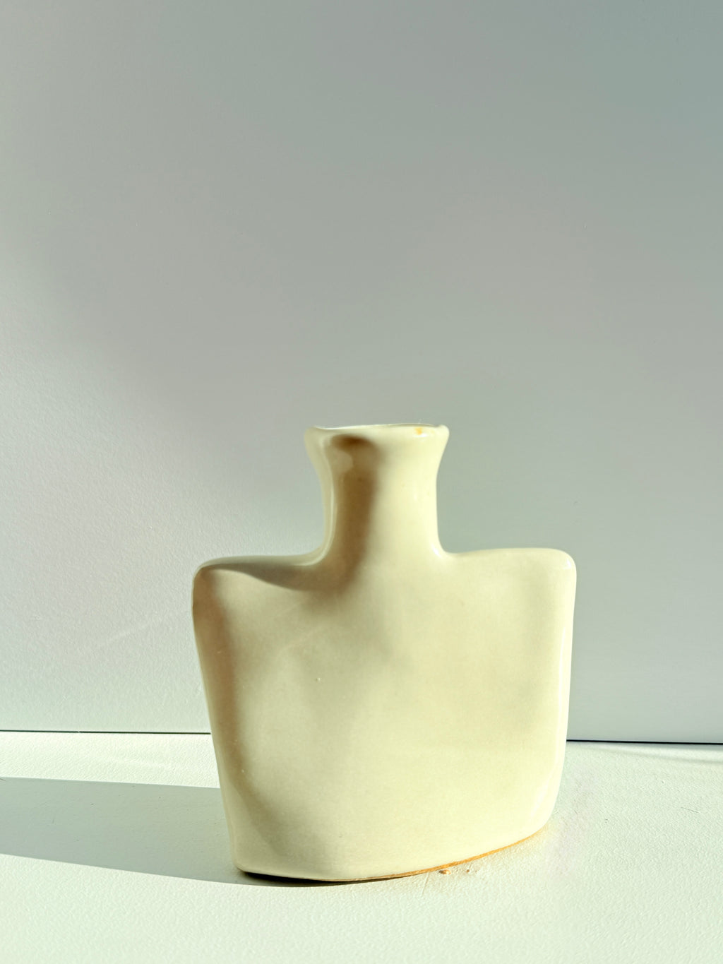 A small sculptural ceramic vase with a matte off-white finish, featuring a wide rounded base and a narrow neck, set against a minimalist light gray background.