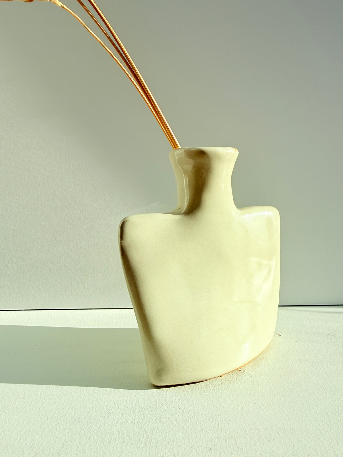 A cream-colored ceramic vase with a unique rounded, organic shape and a short neck, holding two thin copper reeds, set against a minimalist light gray background.