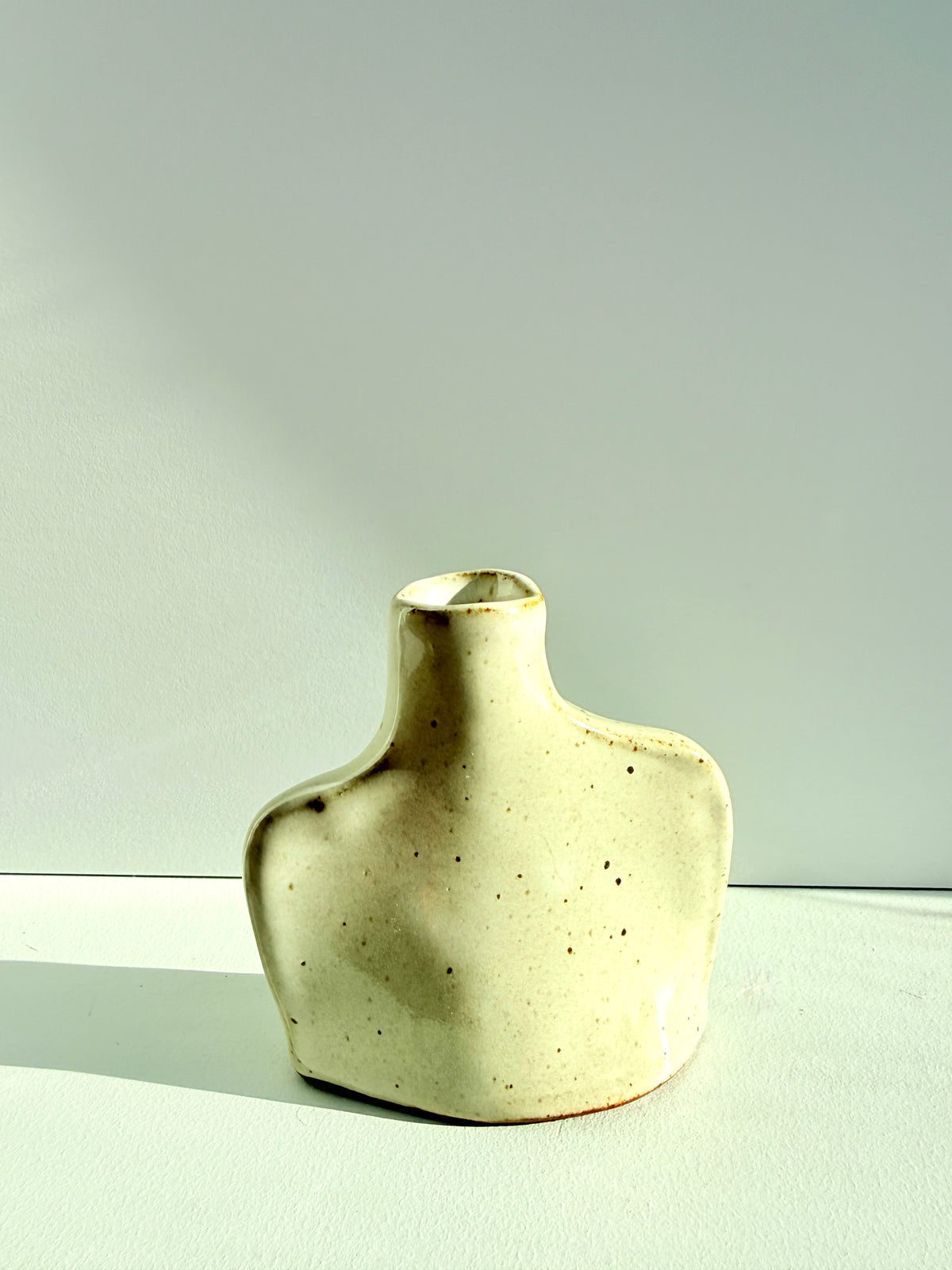 A small sculptural ceramic vase with a wide, rounded base and a narrow neck, featuring a speckled cream glaze with subtle brown speckles, set against a minimalist light gray background.