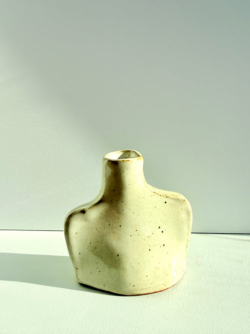 A small sculptural ceramic vase with a wide, rounded base and a narrow neck, featuring a speckled cream glaze with subtle brown speckles, set against a minimalist light gray background.