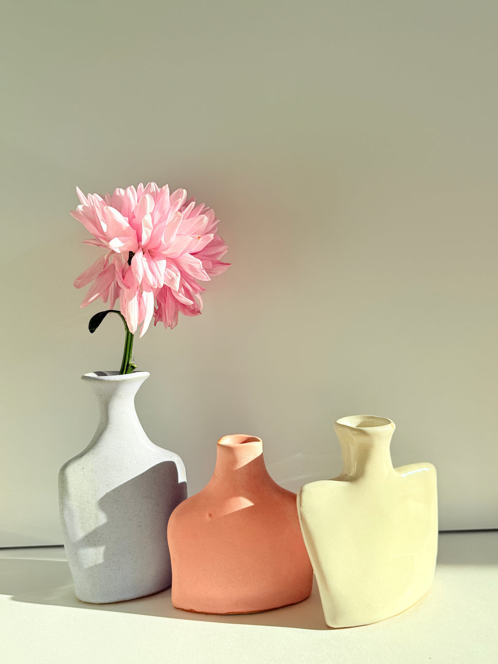 Three minimalist ceramic vases in white, terracotta, and cream, with a single pink flower in the left vase, set against a neutral beige background.