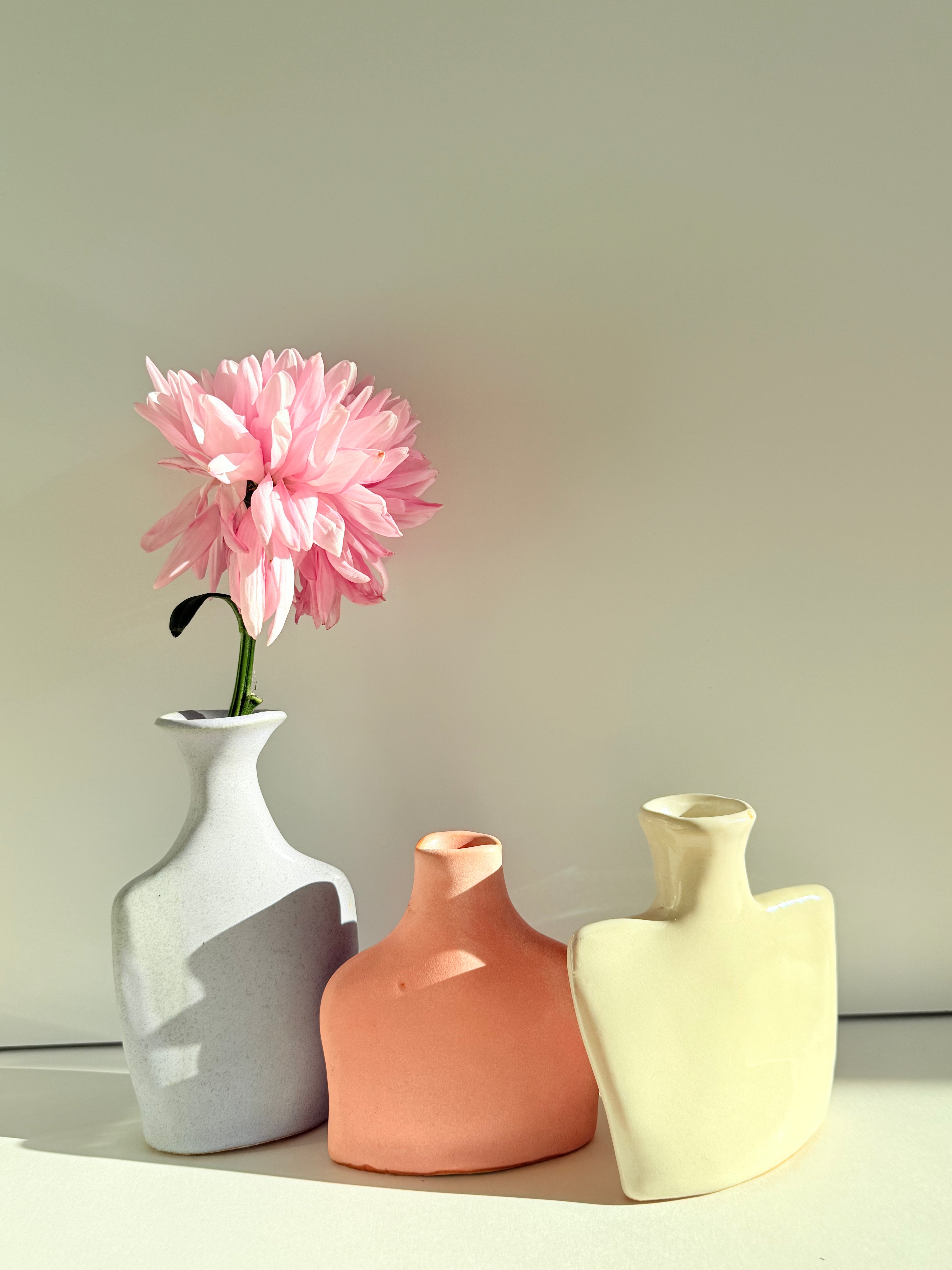 Three minimalist ceramic vases in white, terracotta, and cream, with a single pink flower in the left vase, set against a neutral beige background.