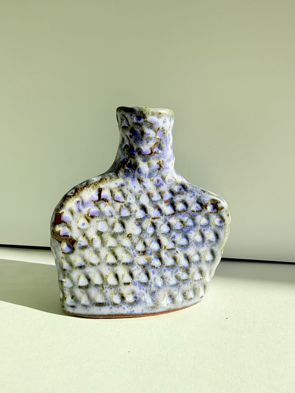 A small handmade ceramic bottle with a textured, dimpled surface and a narrow neck, decorated with a white and light purple glaze accented by brown speckles, set against a plain light green background.