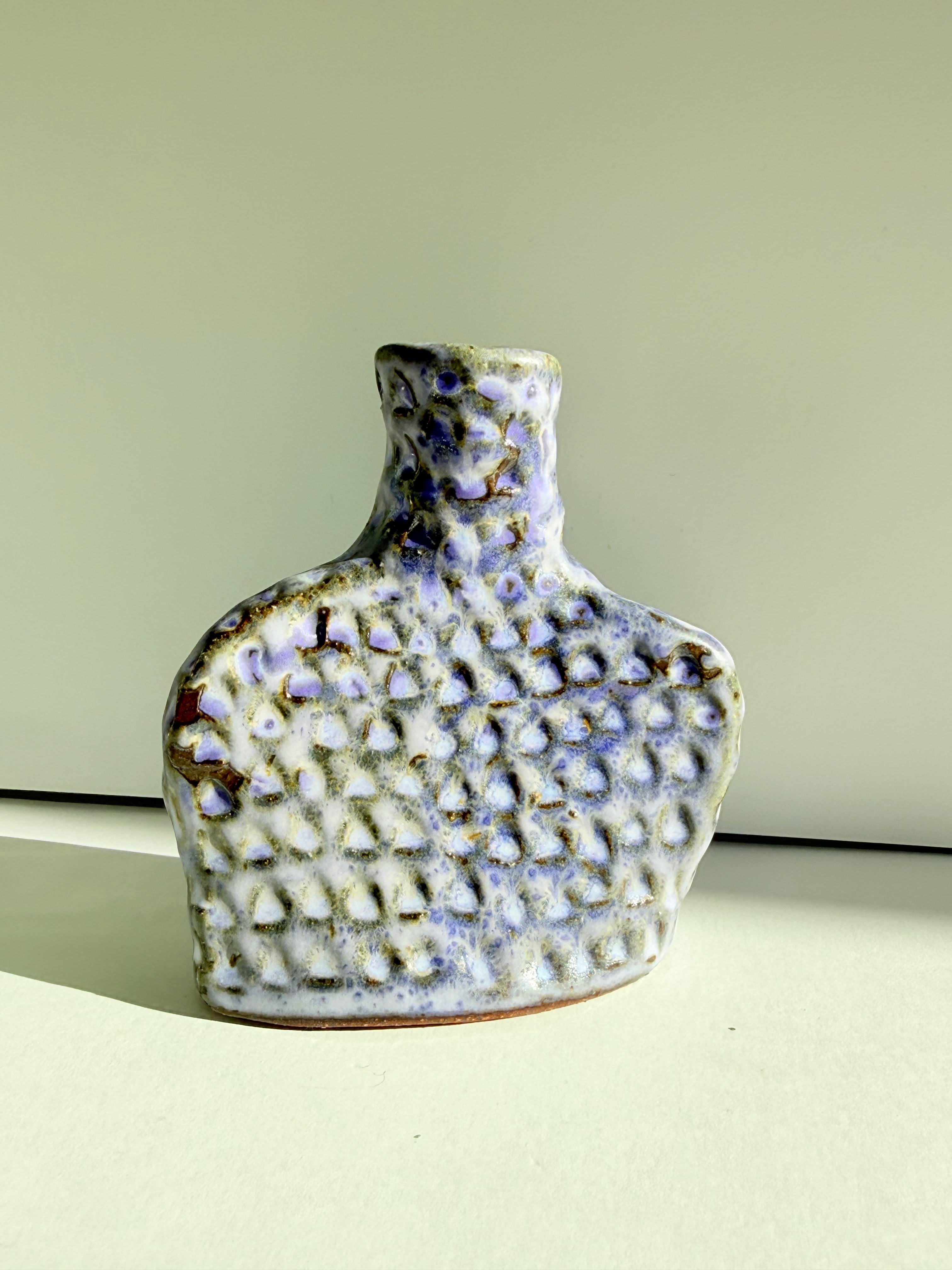 A small handmade ceramic bottle with a textured, dimpled surface and a narrow neck, decorated with a white and light purple glaze accented by brown speckles, set against a plain light green background.