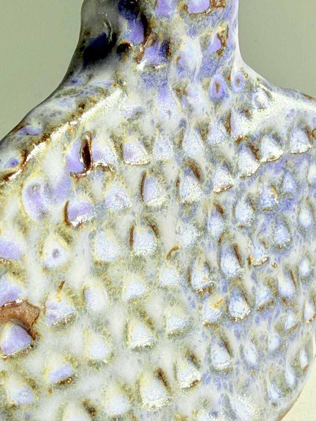 Close-up of a textured ceramic surface with a glossy white and lavender-purple glaze, featuring raised organic bumps outlined in metallic gold and brown.