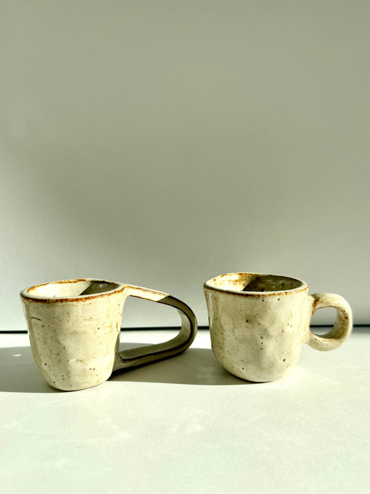 Two handmade ceramic mugs with textured off-white glazes and earthy brown speckles, one featuring a unique integrated handle design, set against a neutral light gray background.