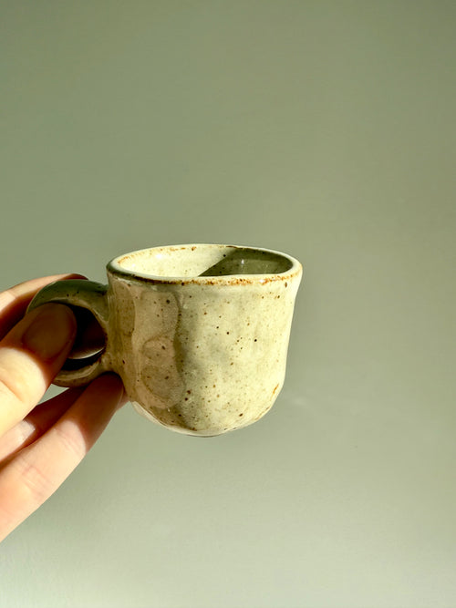A handmade ceramic coffee cup with a rustic handle, held by a hand against a plain light gray background.