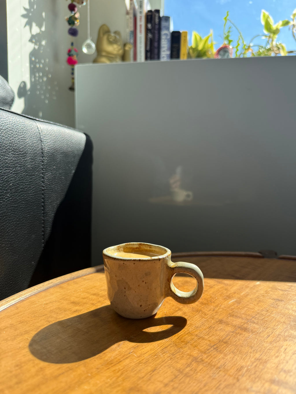 A small handmade ceramic coffee mug with a speckled beige and brown glaze sits on a wooden table in warm sunlight, with a dark leather chair on the left and a modern gray wall in the background.