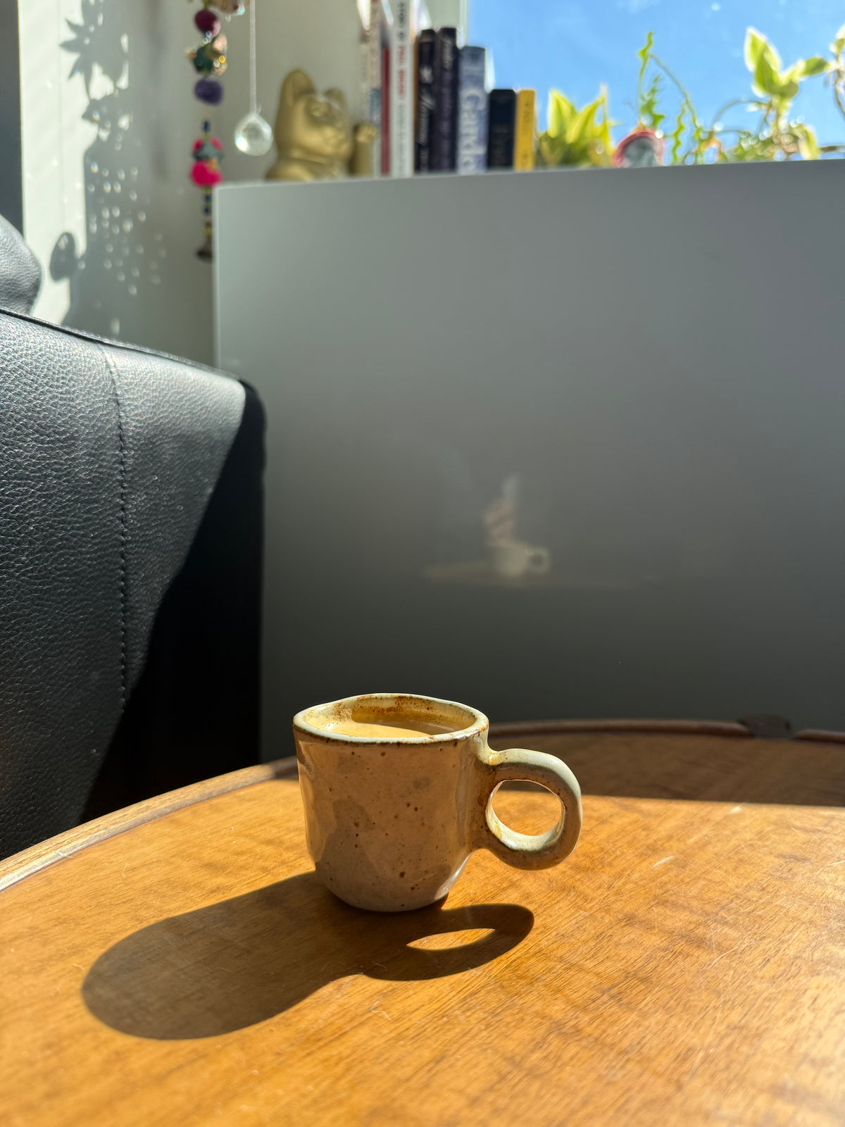 A small handmade ceramic coffee mug with a speckled beige and brown glaze sits on a wooden table in warm sunlight, with a dark leather chair on the left and a modern gray wall in the background.