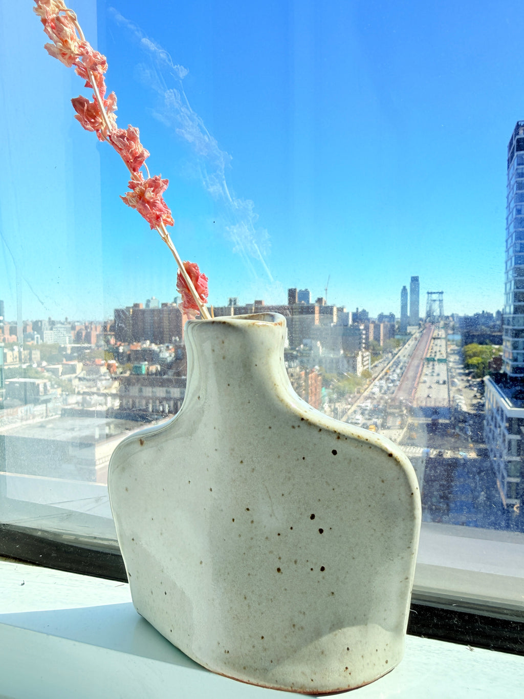 A sculptural ceramic vase with a textured off-white glaze sits on a windowsill, holding a dried pinkish-red flower stem, with a bright city skyline and blue sky visible through the window behind it.