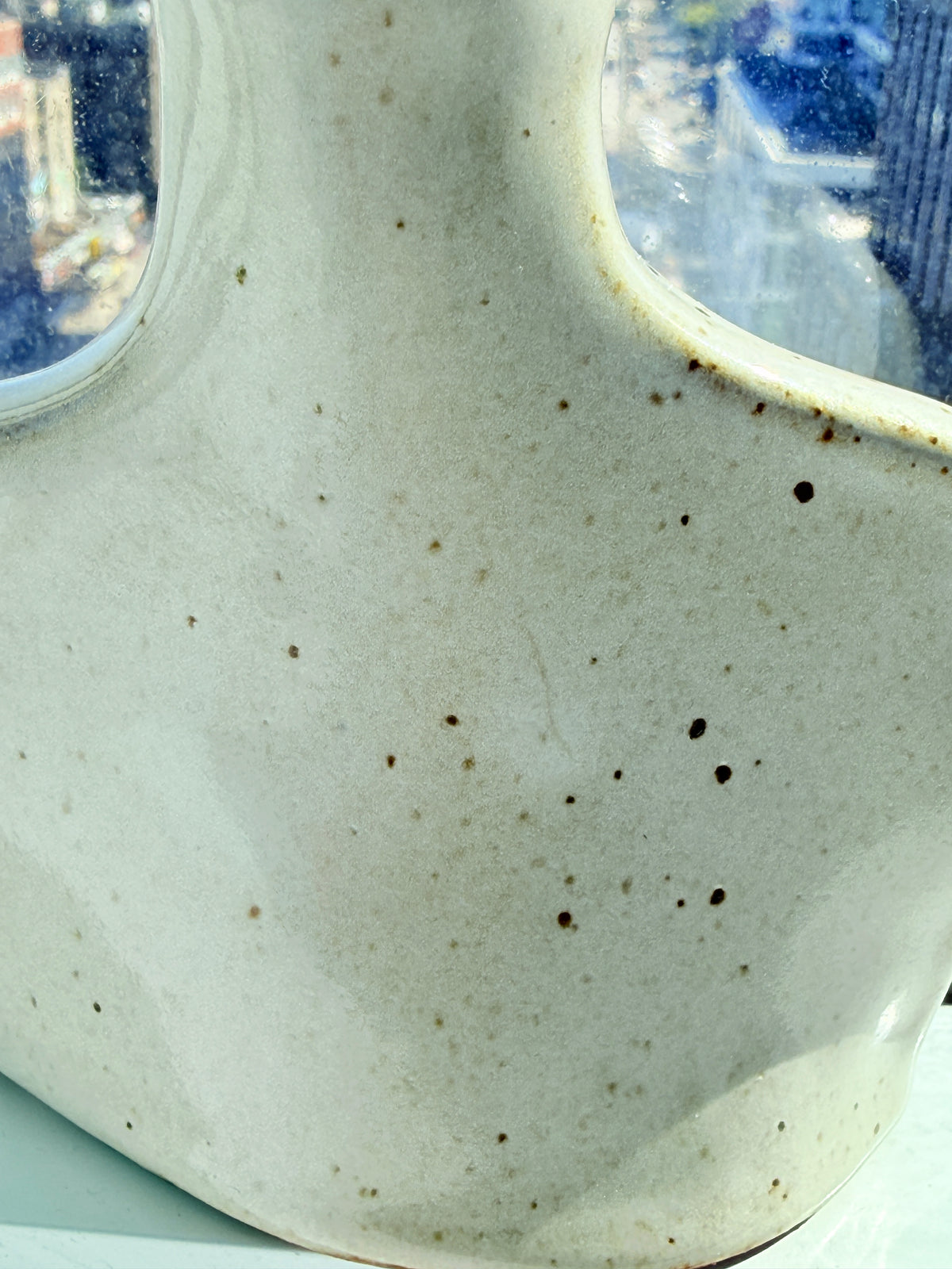 Close-up of a ceramic vessel with a speckled off-white glaze and dark brown spots, showing a curved rim and reflective window background.