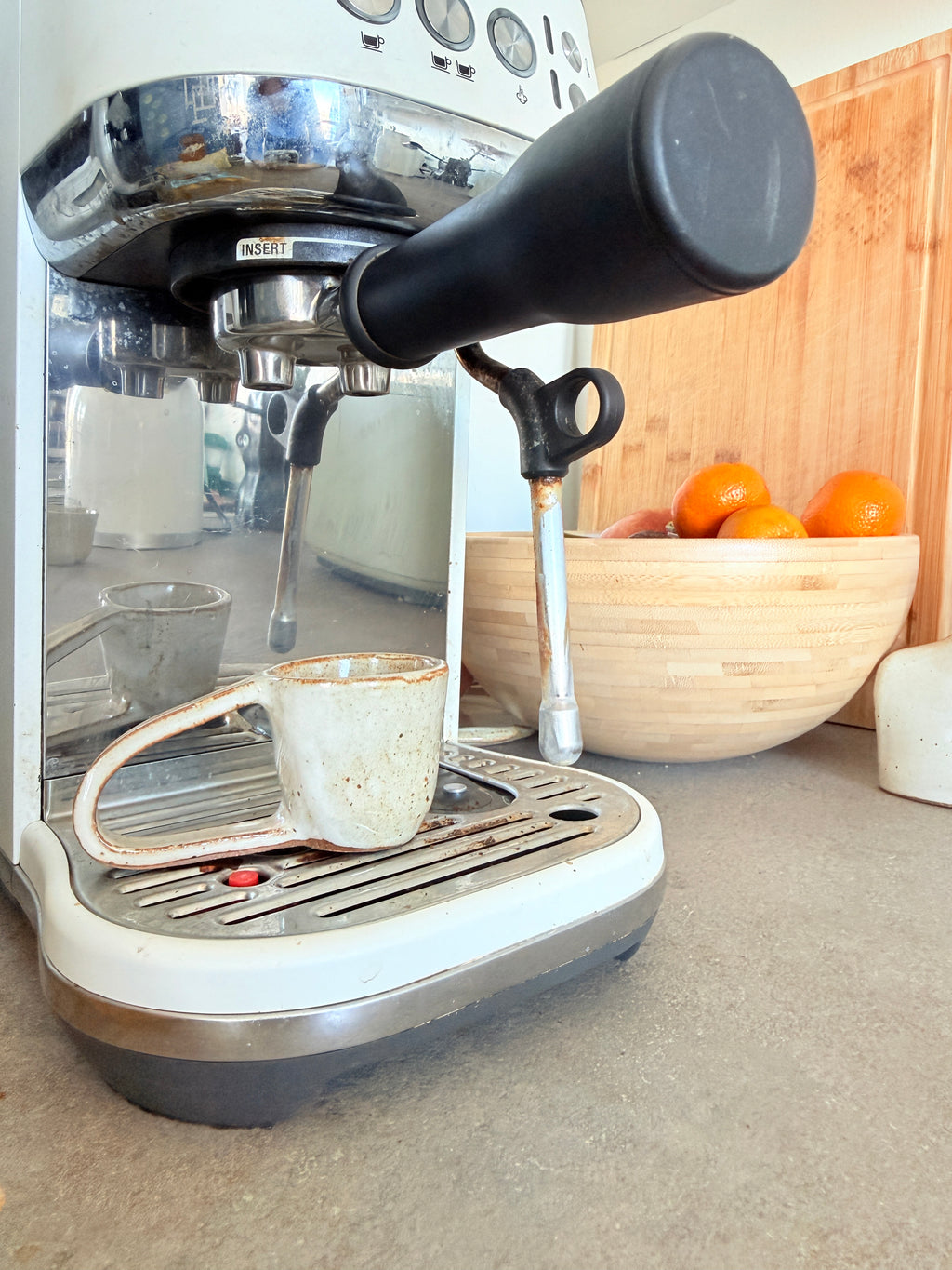 A close-up of a white and stainless steel espresso machine with a black steam wand handle, shown with a ceramic mug on its drip tray in a kitchen setting. A wooden bowl containing oranges sits in the background.
