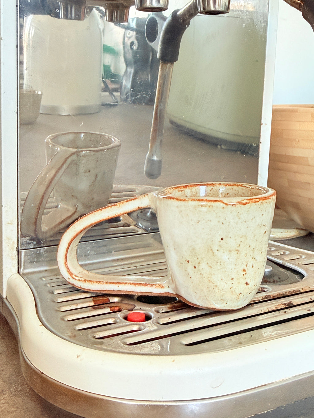 A handmade ceramic coffee mug with a speckled cream glaze and a rustic orange-brown rim, sitting on the drip tray of a coffee machine.