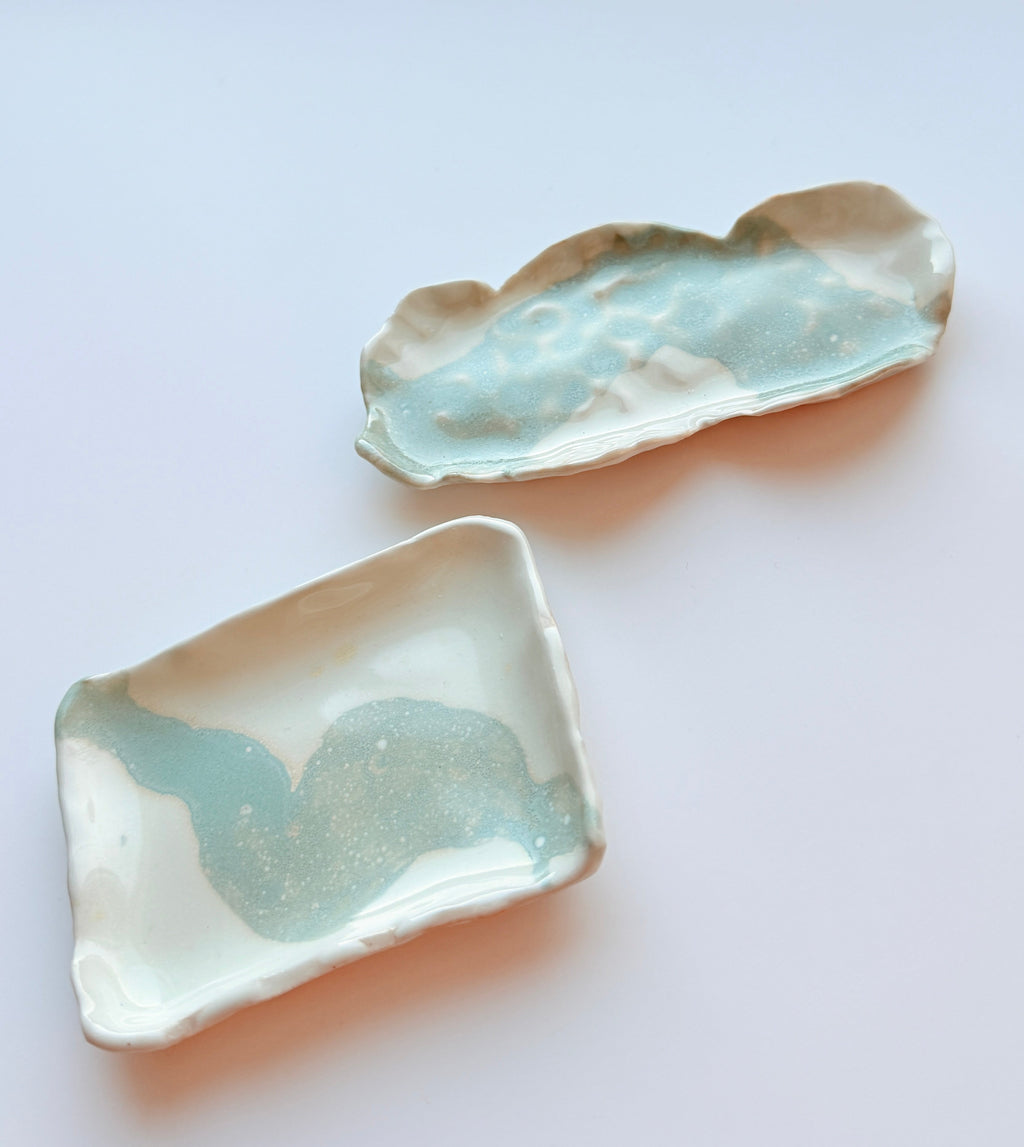 Two small handmade ceramic trays with organic, cloud-like shapes and a glossy light blue-green glaze, displayed on a clean white surface.