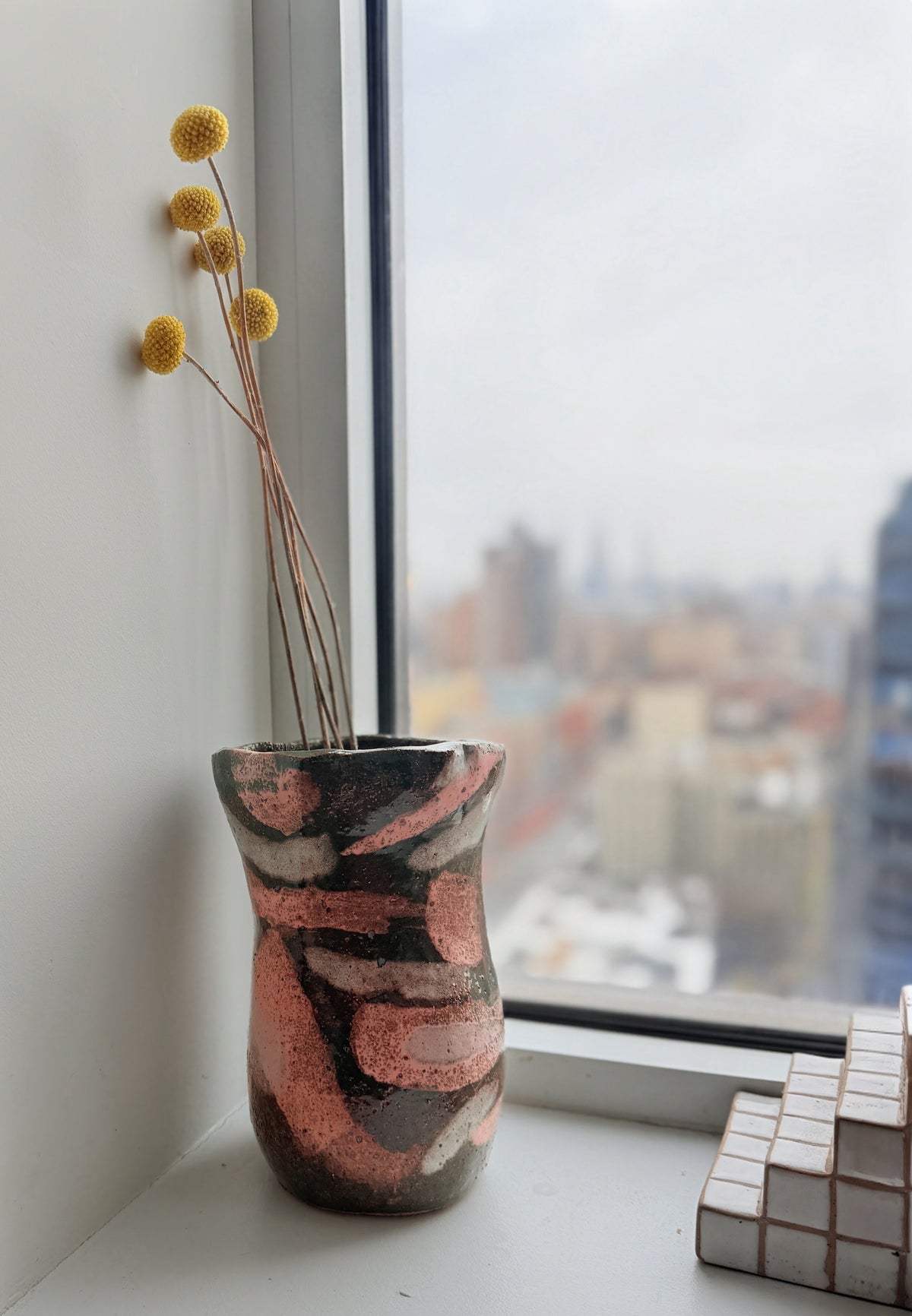 A handmade ceramic vase with a wavy, ribbed design and a speckled pattern of terracotta pink, dark brown, and cream, holding dried yellow globe flowers, set on a windowsill with a city view outside.