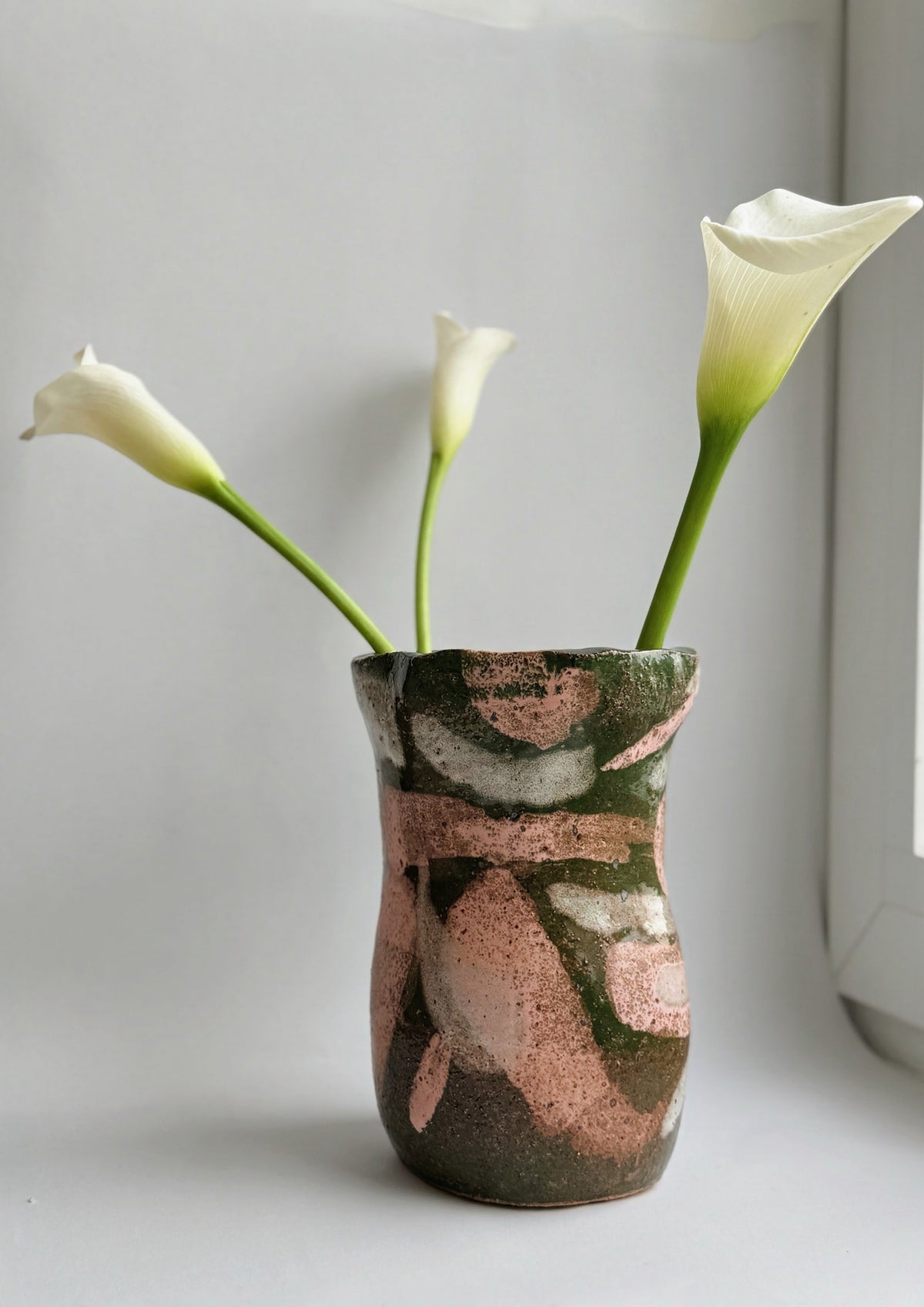 A handmade ceramic vase with a textured, abstract pattern of green, brown, and metallic rose gold, holding three white calla lilies against a neutral light gray background.