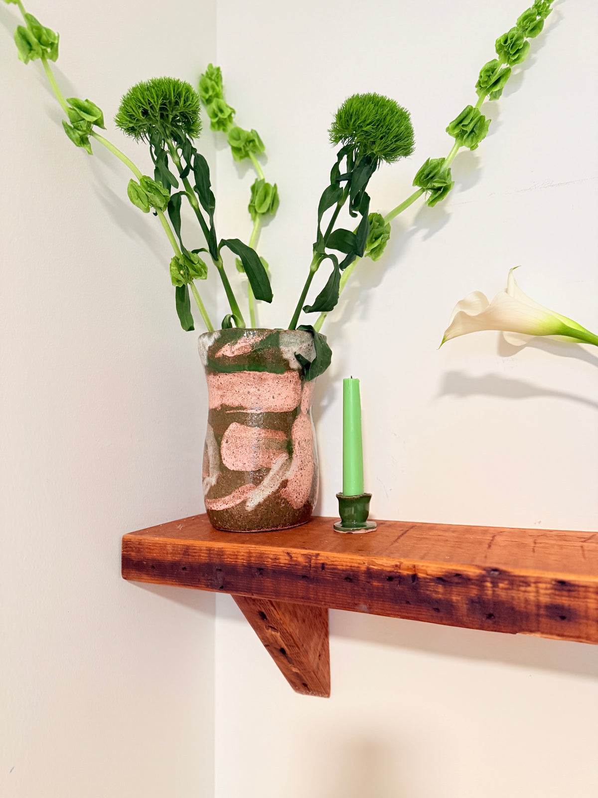 A rustic ceramic vase with a pink, green, and brown abstract pattern, holding green flower stems, displayed on a wooden floating shelf next to a green candle.