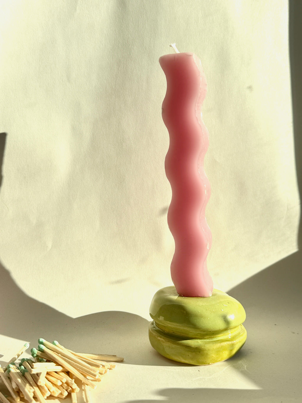 Pink wavy candle on a green base with matches beside it against a light curtain background