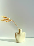 A minimalist cream-colored ceramic vase with a sculptural, rounded rectangular body and a narrow neck, holding a single sprig of dried wheat against a clean, light gray background.