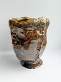 A handmade ceramic cup with an organic, wavy rim and a textured, organic glaze featuring brown, gold, gray, and black tones, set against a plain light gray background.