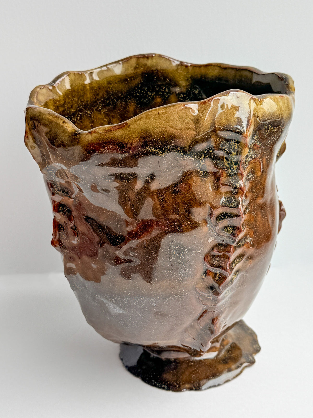 A handmade ceramic bowl with a wavy, organic rim and a small pedestal base, featuring a glossy glaze with rich brown, amber, and dark charcoal tones.