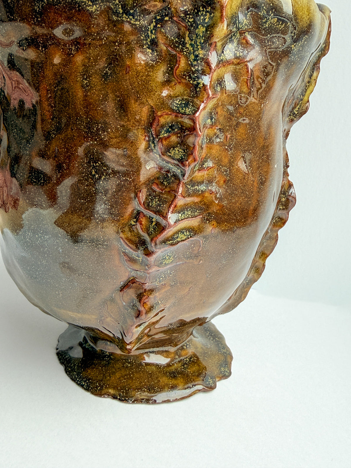 A close-up of a sculptural ceramic vase with a glossy, organic, organic-like glaze in earthy brown, amber, and gold tones with subtle yellow and green marbling.