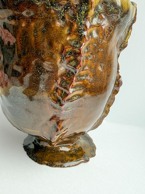 A close-up of a sculptural ceramic vase with a glossy, organic, organic-like glaze in earthy brown, amber, and gold tones with subtle yellow and green marbling.