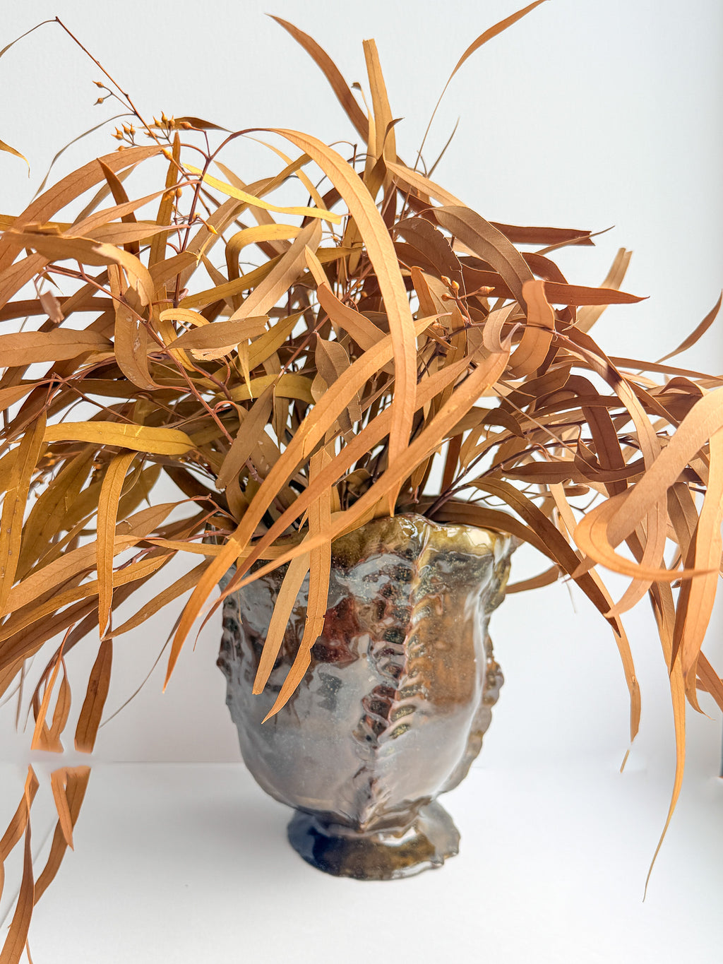A decorative ceramic planter with a textured, metallic bronze and silver finish, filled with dried, long, narrow brown leaves, set against a plain white background.
