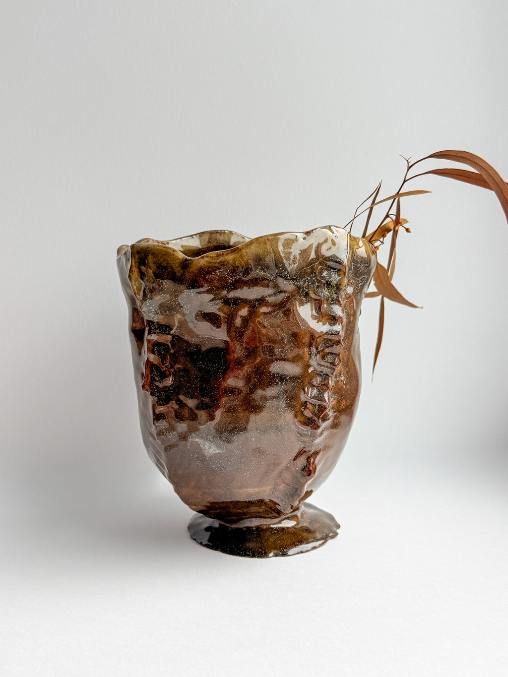 A handmade ceramic bud vase with a glossy, organic brown and amber glaze, styled with a dried leafy branch on a light gray background.