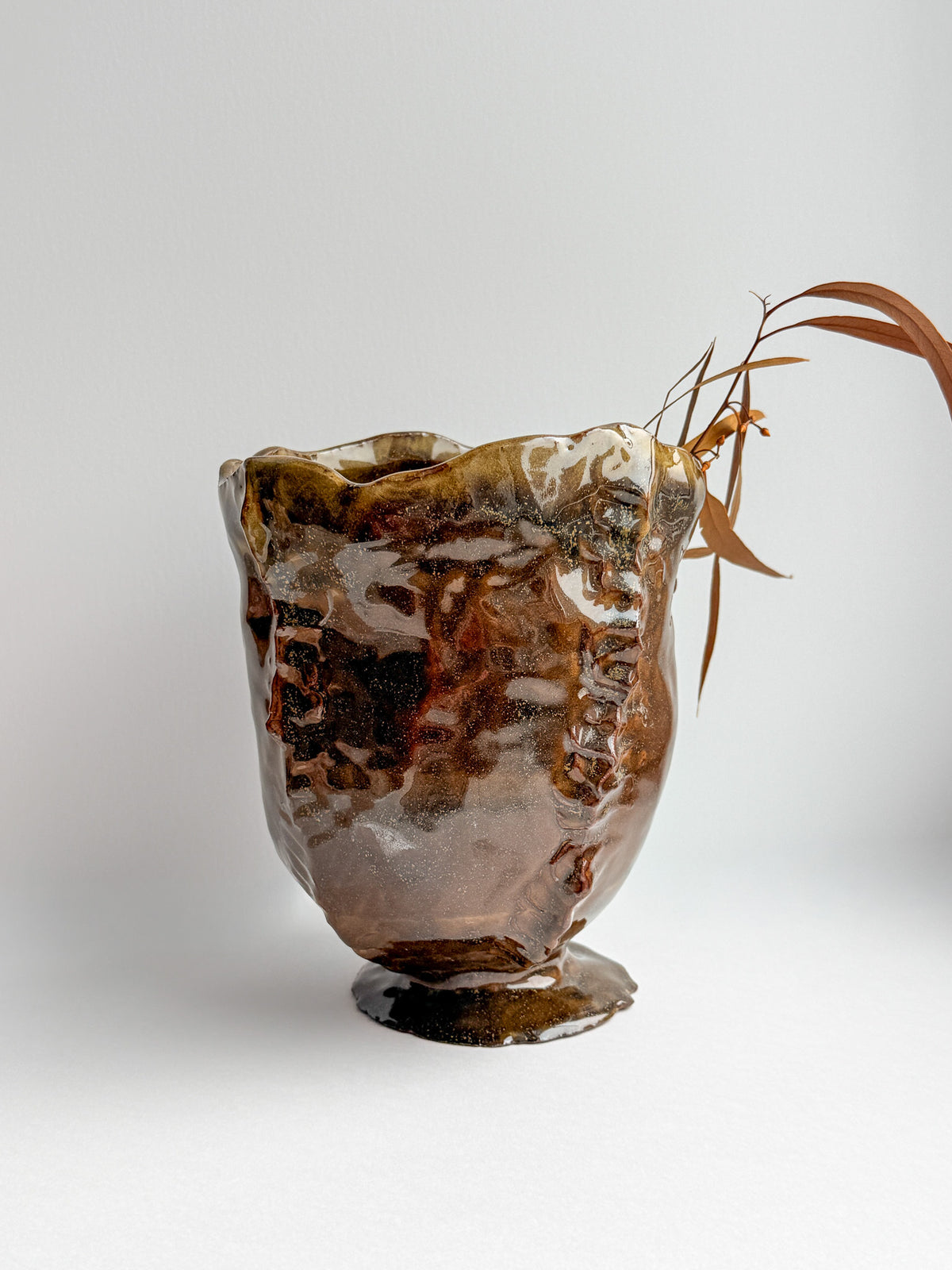 A handmade ceramic bud vase with a glossy, organic brown and amber glaze, styled with a dried leafy branch on a light gray background.