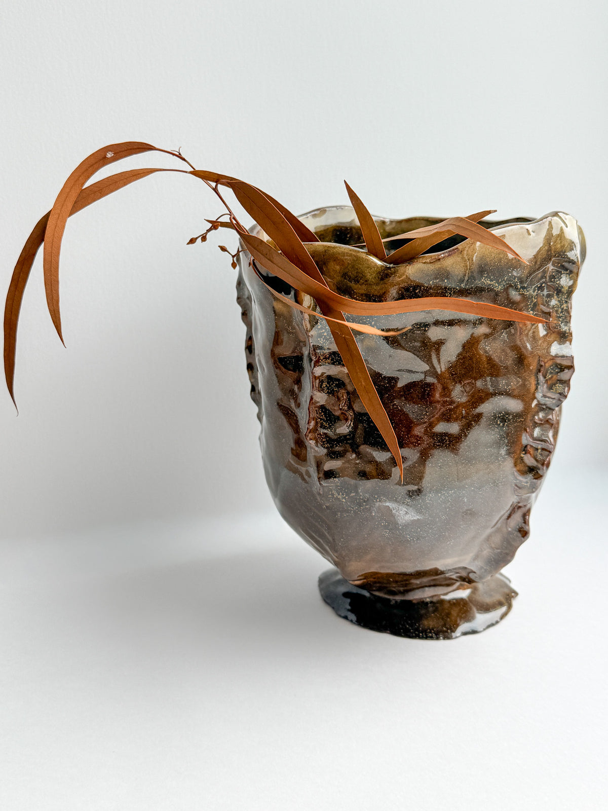A rustic, handcrafted ceramic planter with a textured, organic shape and a glossy brown and gray glaze, styled with dried brown leaves on a clean white background.