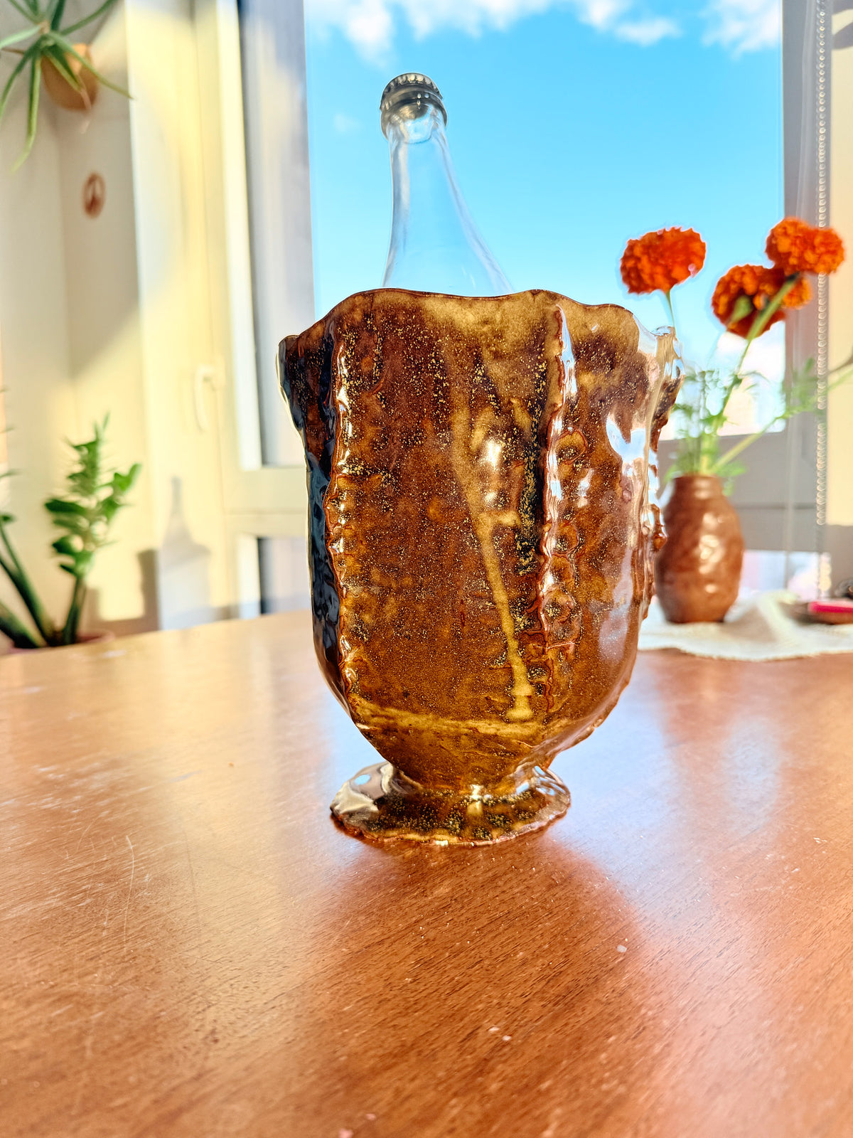A handmade amber-brown textured glass vase with a clear glass bottle inserted inside, displayed on a wooden table in a sunlit room with flowers in the background.