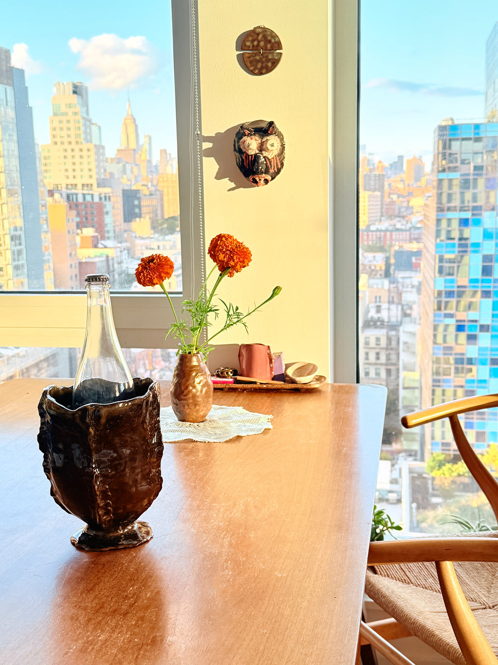 A styled dining table in a bright room with a large window overlooking a city skyline. On the table are a dark textured ceramic vase with a glass bottle, a small brown vase with orange flowers.