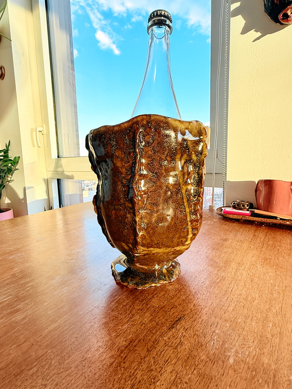 A clear glass bottle with a thick, textured amber-brown base and a black screw cap, standing on a wooden table in a brightly lit room with a window behind it.
