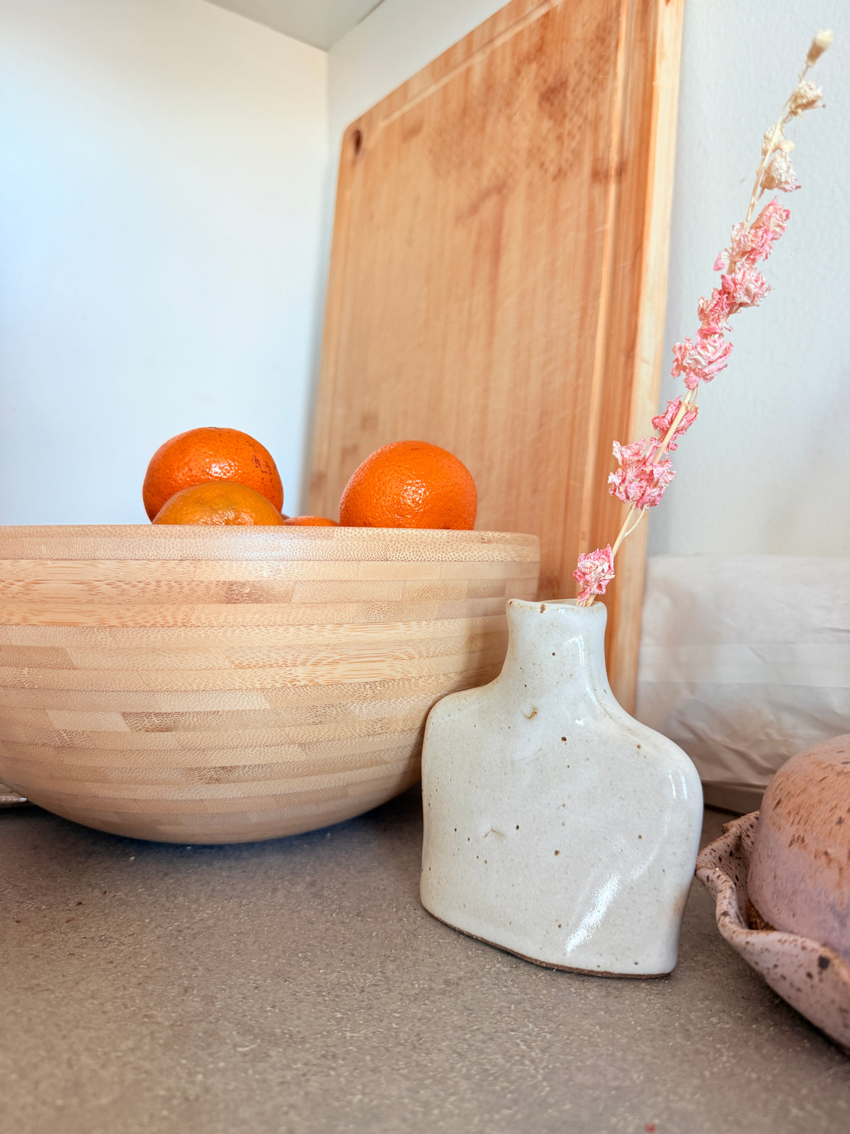 A ceramic vase with a textured, speckled off-white glaze holding a dried pink flower, set next to a large wooden bowl with oranges on a countertop.