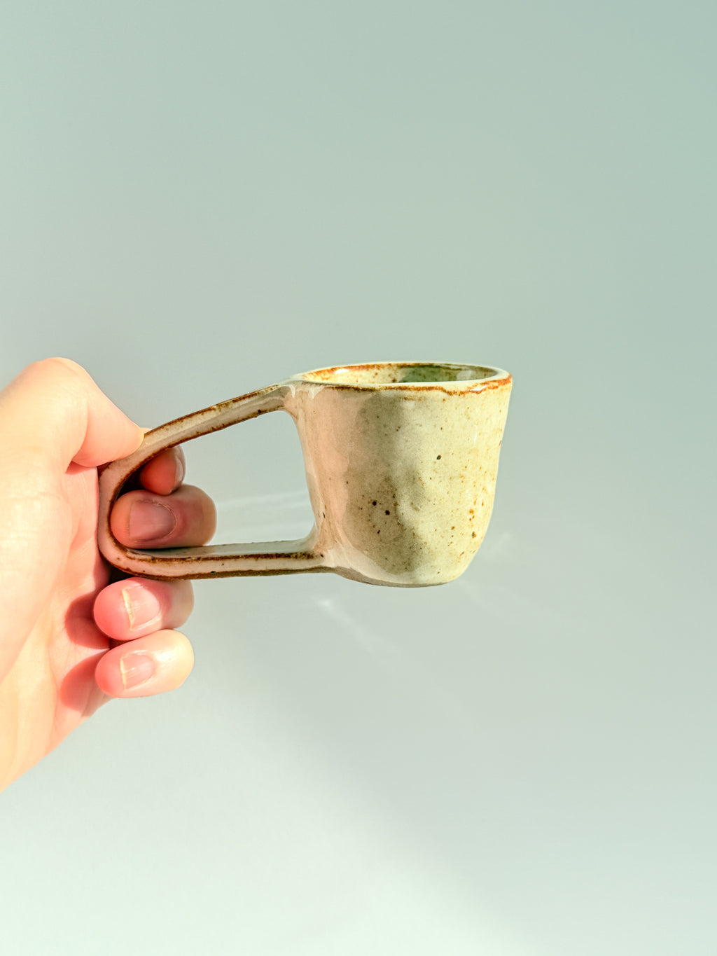 A hand holding a small handmade ceramic mug with a unique loop handle, featuring a textured off-white glaze with brown speckles and a rustic brown rim, set against a solid light gray background.