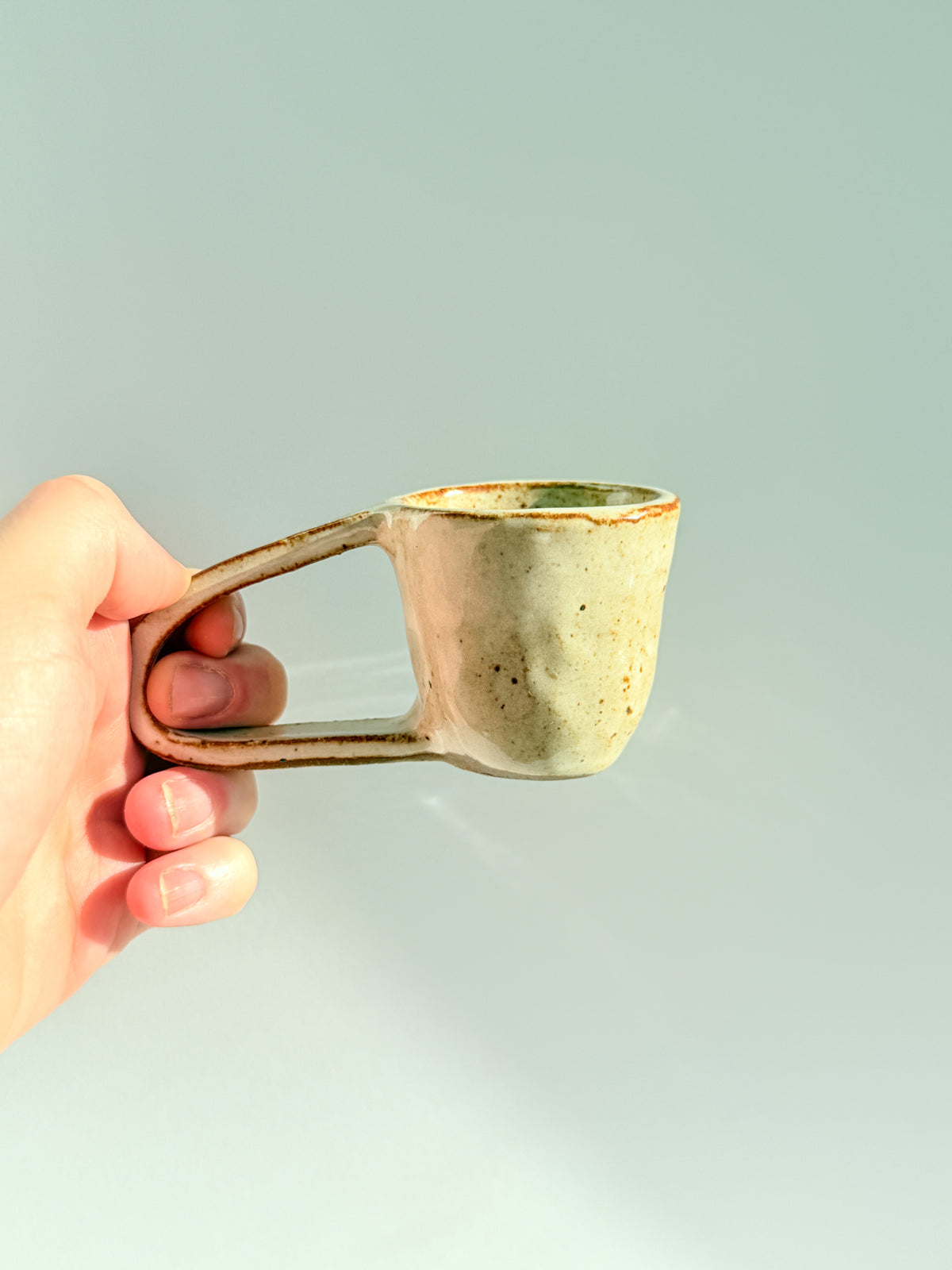 A hand holding a small handmade ceramic mug with a unique loop handle, featuring a textured off-white glaze with brown speckles and a rustic brown rim, set against a solid light gray background.