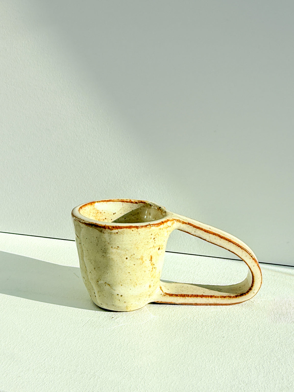 A handmade ceramic mug with a unique loop handle, featuring a textured off-white glaze with brown speckles and a thin terracotta rim, set against a minimalist light gray background.