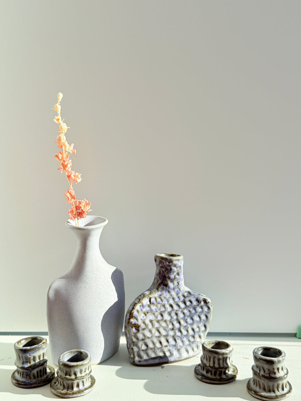 A set of handmade ceramic vases and candle holders displayed on a white surface against a light gray background. The set includes a tall matte white vase with dried coral flowers, a textured gray and white bottle-shaped vase, and four small textured gray candle cups.