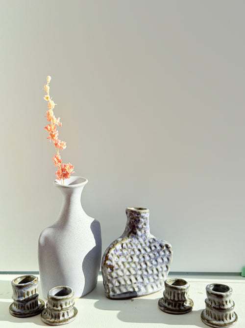 A set of handmade ceramic vases and candle holders displayed on a white surface against a light gray background. The set includes a tall matte white vase with dried coral flowers, a textured gray and white bottle-shaped vase, and four small textured gray candle cups.