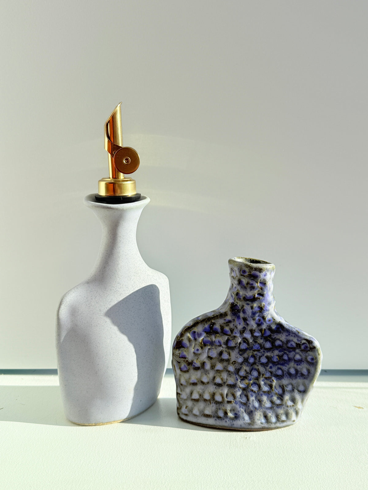 Two decorative ceramic bottles on a white surface against a light gray background. The left bottle is matte off-white with a gold metal pour spout.