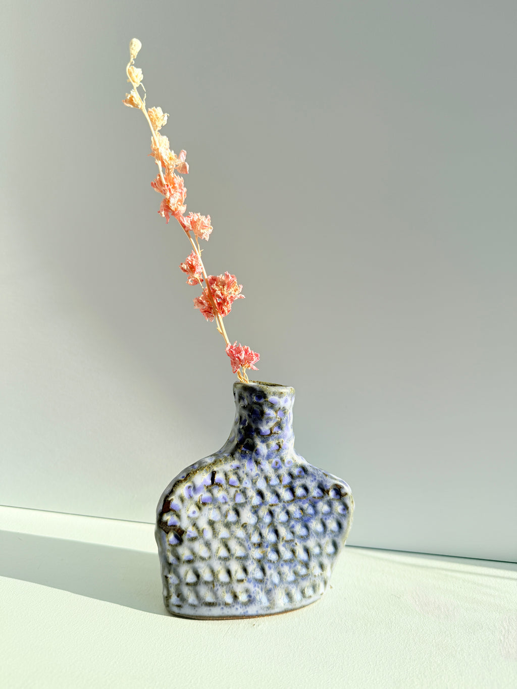 A textured ceramic vase with a blue, white, and black glaze, holding a dried pinkish-orange flower stem, set against a neutral gray background.