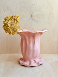 Pink ceramic vase with a textured surface holding a dried yellow flower against a light wooden background