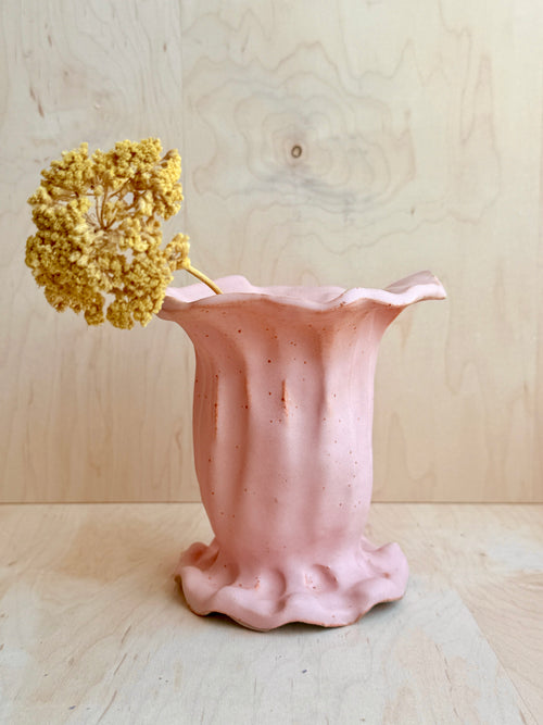 Pink ceramic vase with a textured surface holding a dried yellow flower against a light wooden background